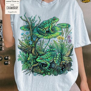 May include: Light blue Comfort Colors tee featuring a detailed illustration of two green frogs in a pond setting. The design includes lily pads, flowers, and aquatic plants. The shirt has the text "CHAMBRAY" and "Comfort Colors Tee".