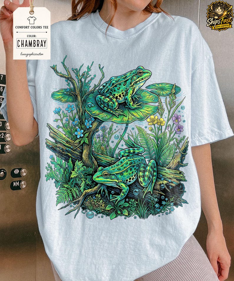 May include: Light blue Comfort Colors tee featuring a detailed illustration of two green frogs in a pond setting. The design includes lily pads, flowers, and aquatic plants. The shirt has the text "CHAMBRAY" and "Comfort Colors Tee".