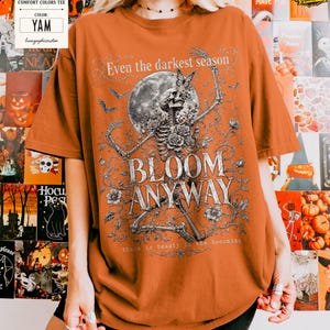 May include: A yam-colored t-shirt featuring a graphic print of a skeleton surrounded by flowers, a butterfly, and a full moon. The text reads: Even the darkest season, BLOOM ANYWAY, and there is beauty in the becoming. The shirt has short sleeves.