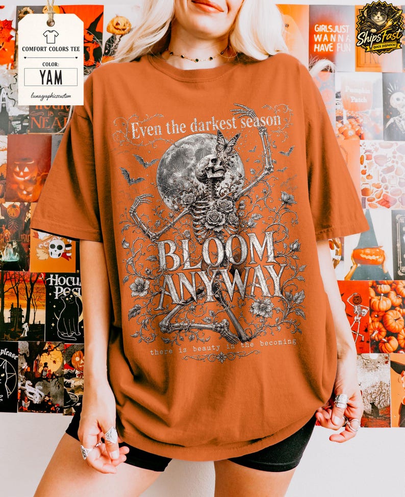 May include: A yam-colored t-shirt featuring a graphic print of a skeleton surrounded by flowers, a butterfly, and a full moon. The text reads: Even the darkest season, BLOOM ANYWAY, and there is beauty in the becoming. The shirt has short sleeves.