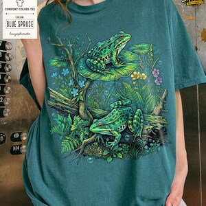 May include: Blue Spruce Comfort Colors tee featuring a detailed illustration of two green frogs amidst aquatic plants, flowers, and bubbles. The design includes a log and lily pads, creating a serene, nature-inspired scene. The shirt has a tag that says "Comfort Colors Tee" and "Blue Spruce".