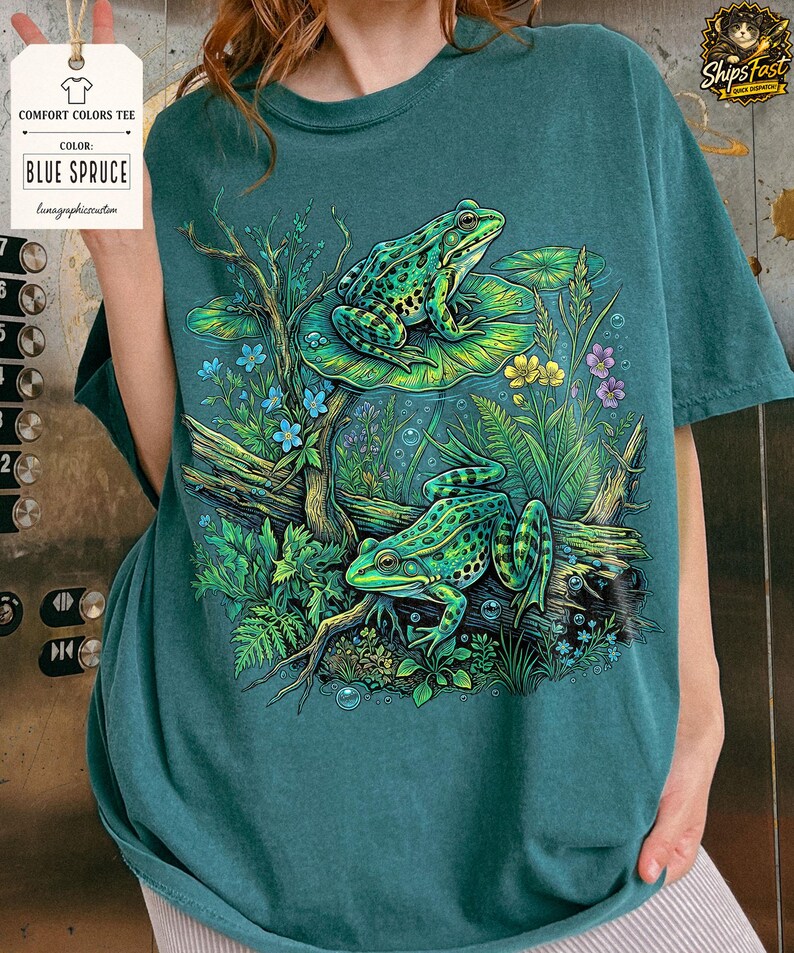May include: Blue Spruce Comfort Colors tee featuring a detailed illustration of two green frogs amidst aquatic plants, flowers, and bubbles. The design includes a log and lily pads, creating a serene, nature-inspired scene. The shirt has a tag that says "Comfort Colors Tee" and "Blue Spruce".