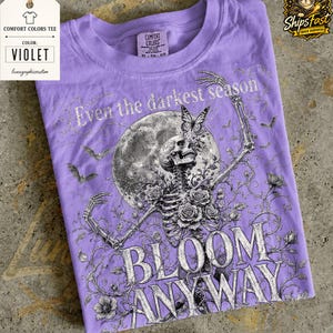 May include: A violet t-shirt featuring a graphic print of a skeleton surrounded by roses, a large moon, bats, and a butterfly. The text reads Even the darkest season BLOOM ANYWAY. The shirt has a crew neckline and a Comfort Colors brand label inside.