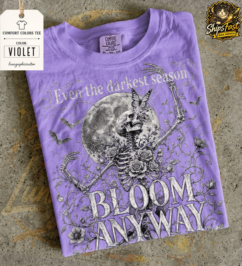 May include: A violet t-shirt featuring a graphic print of a skeleton surrounded by roses, a large moon, bats, and a butterfly. The text reads Even the darkest season BLOOM ANYWAY. The shirt has a crew neckline and a Comfort Colors brand label inside.
