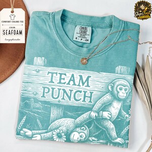 May include: Seafoam green Comfort Colors tee featuring a monkey graphic and the text "TEAM PUNCH". The t-shirt has a crew neck and short sleeves. A gold necklace is draped across the shirt.