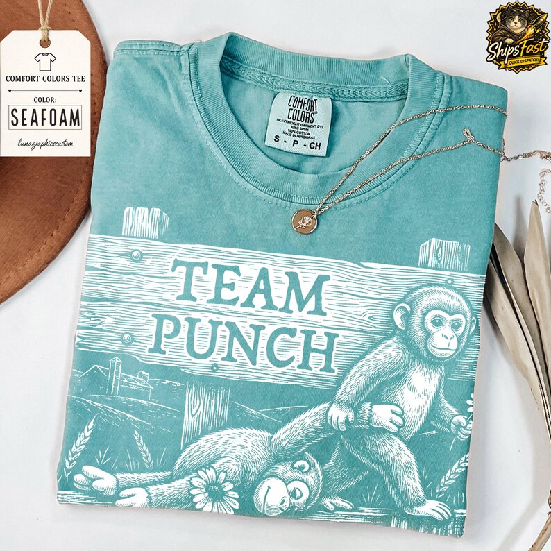 May include: Seafoam green Comfort Colors tee featuring a monkey graphic and the text "TEAM PUNCH". The t-shirt has a crew neck and short sleeves. A gold necklace is draped across the shirt.