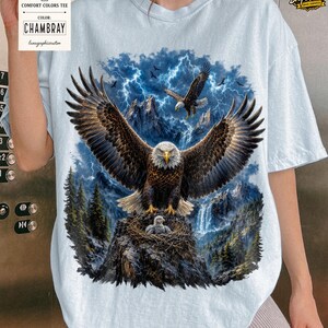 May include: Light blue Comfort Colors tee featuring a detailed graphic of an eagle with outstretched wings, a nest with eaglets, and a mountain backdrop with lightning. The shirt includes the color "Chambray" on the tag.