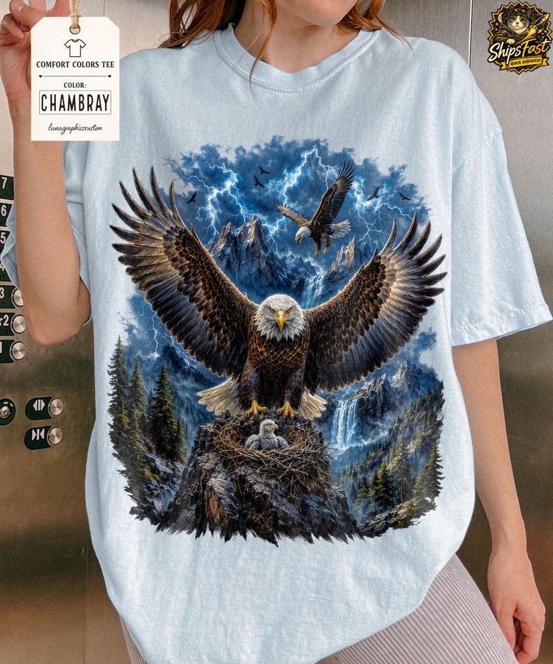 May include: Light blue Comfort Colors tee featuring a detailed graphic of an eagle with outstretched wings, a nest with eaglets, and a mountain backdrop with lightning. The shirt includes the color "Chambray" on the tag.