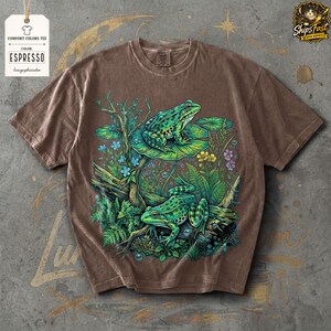 May include: Espresso-colored Comfort Colors tee featuring a detailed illustration of two green frogs in a pond setting with lily pads, plants, and flowers. The shirt includes the text "COMFORT COLORS TEE" and "ESPRESSO".