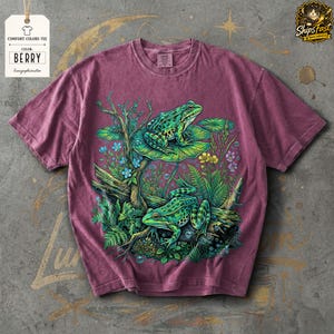 May include: Berry-colored Comfort Colors tee featuring a detailed graphic of two green frogs amidst aquatic plants and flowers. The design includes lily pads, branches, and small blue and yellow flowers. The shirt has a tag that says "Comfort Colors Tee" and "Color: Berry".
