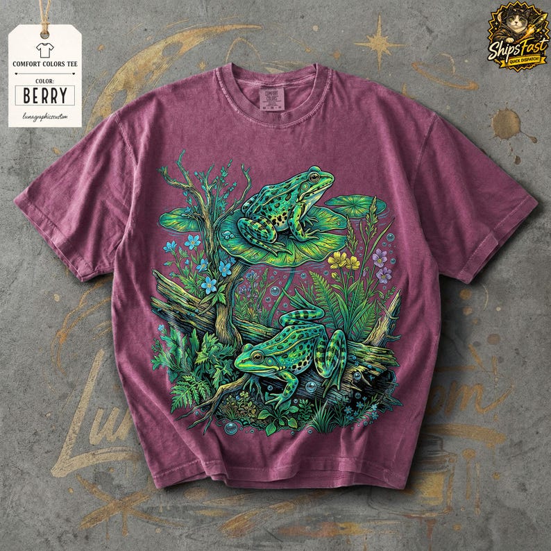 May include: Berry-colored Comfort Colors tee featuring a detailed graphic of two green frogs amidst aquatic plants and flowers. The design includes lily pads, branches, and small blue and yellow flowers. The shirt has a tag that says "Comfort Colors Tee" and "Color: Berry".