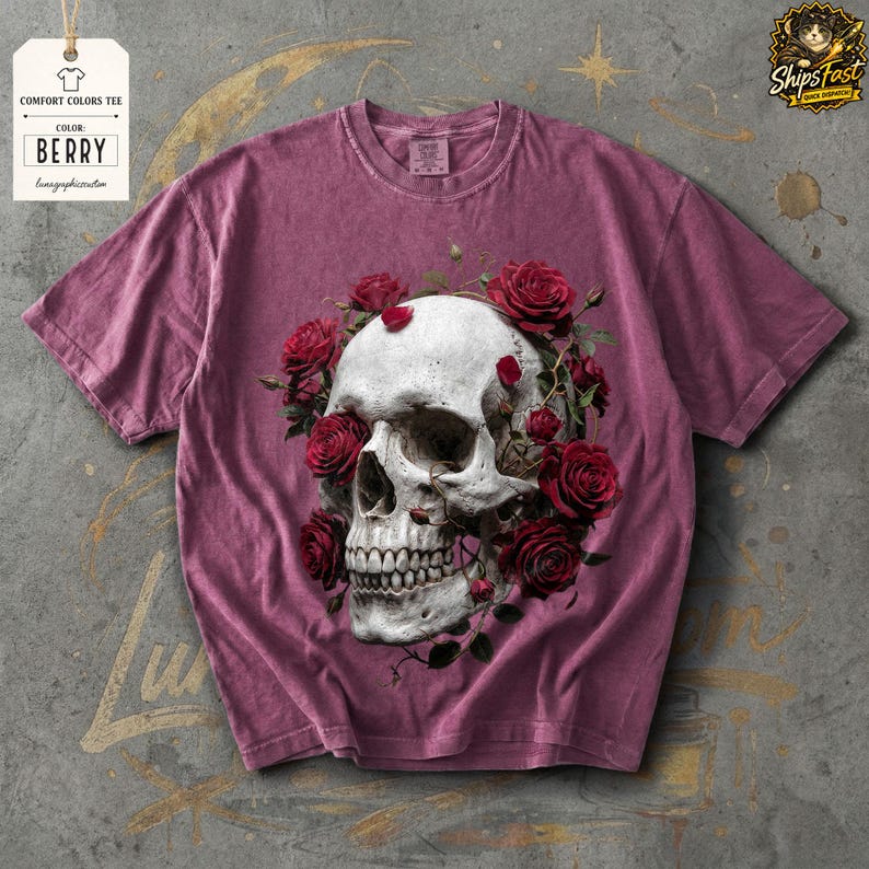 May include: Berry-colored comfort colors tee featuring a detailed skull design encircled by red roses. The shirt has a crew neck and short sleeves. The tag reads "Comfort Colors Tee" and "Berry".