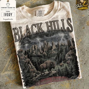 May include: Ivory Comfort Colors tee featuring a detailed graphic of the Black Hills with bison, trees, and lightning. The text "BLACK HILLS" arches across the top, with "Outdoor Discovery" at the bottom.