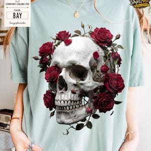 May include: A light blue Comfort Colors tee shirt featuring a detailed graphic of a human skull adorned with vibrant red roses. The shirt's color is "Bay" as indicated on the tag.