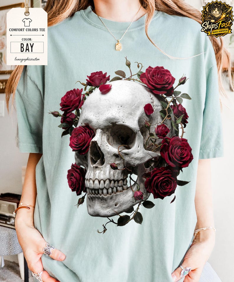 May include: A light blue Comfort Colors tee shirt featuring a detailed graphic of a human skull adorned with vibrant red roses. The shirt's color is "Bay" as indicated on the tag.