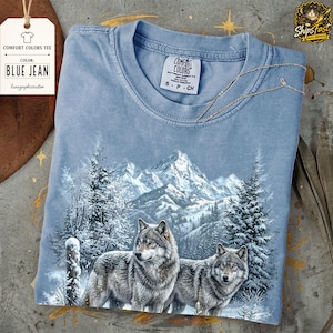 Pode incluir: Camiseta Comfort Colors azul jeans com um gráfico detalhado de dois lobos em uma paisagem montanhosa nevada. A camiseta inclui uma etiqueta com o nome da cor e informações da marca. A camiseta é tamanho P.