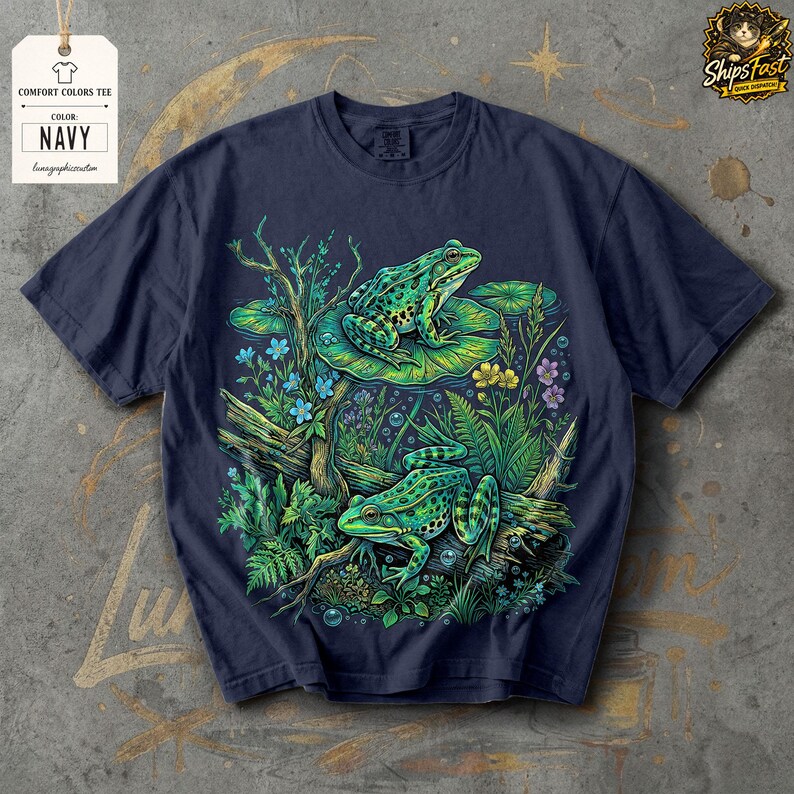May include: Navy blue Comfort Colors tee featuring a detailed illustration of two green frogs in a pond setting. The design includes lily pads, flowers, and aquatic plants. The tag reads "COMFORT COLORS TEE" and "NAVY".