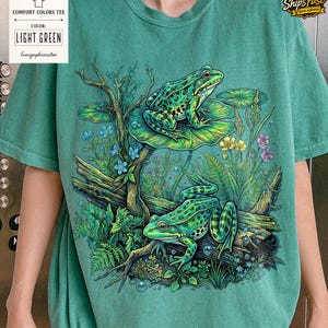 May include: Light green Comfort Colors tee featuring a detailed illustration of two green frogs in a pond setting. The frogs are on a log and lily pads, surrounded by plants and flowers. The shirt has the text "LIGHT GREEN".
