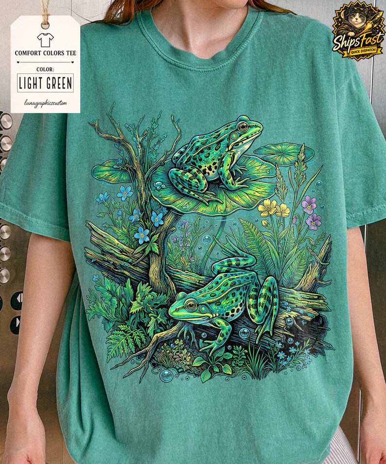 May include: Light green Comfort Colors tee featuring a detailed illustration of two green frogs in a pond setting. The frogs are on a log and lily pads, surrounded by plants and flowers. The shirt has the text "LIGHT GREEN".