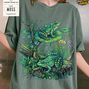 May include: Moss green Comfort Colors tee featuring a detailed illustration of two green frogs amidst aquatic plants and flowers. The frogs are on a lily pad and a log. The shirt has the text "Comfort Colors Tee" and "Moss".