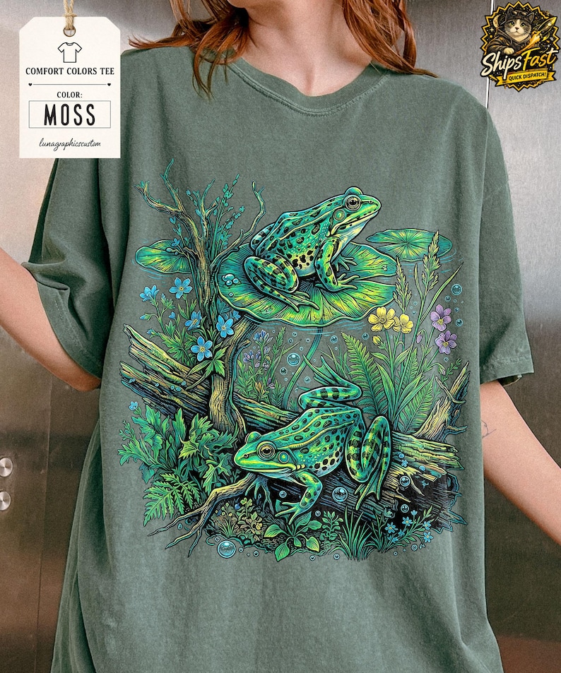 May include: Moss green Comfort Colors tee featuring a detailed illustration of two green frogs amidst aquatic plants and flowers. The frogs are on a lily pad and a log. The shirt has the text "Comfort Colors Tee" and "Moss".