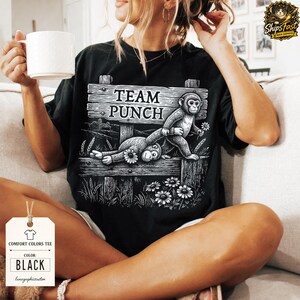 May include: Black Comfort Colors tee with a graphic of two monkeys and the text "TEAM PUNCH" on a wooden sign. The design includes daisies and wheat stalks. The shirt has a tag that says "COMFORT COLORS TEE" and "BLACK".