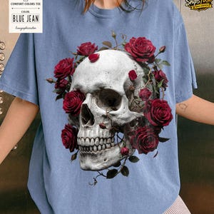 May include: Blue jean Comfort Colors tee featuring a detailed skull design encircled by vibrant red roses. The graphic tee showcases a gothic aesthetic, blending floral and macabre elements. The shirt's color is indicated by a tag that reads "BLUE JEAN".
