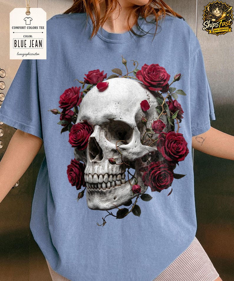 May include: Blue jean Comfort Colors tee featuring a detailed skull design encircled by vibrant red roses. The graphic tee showcases a gothic aesthetic, blending floral and macabre elements. The shirt's color is indicated by a tag that reads "BLUE JEAN".