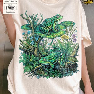 May include: Ivory Comfort Colors tee featuring a vibrant illustration of two green frogs in a pond setting. The design includes lily pads, aquatic plants, and the text "Comfort Colors Tee" and "Ivory".