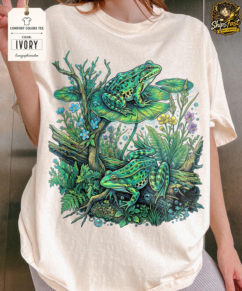 May include: Ivory Comfort Colors tee featuring a vibrant illustration of two green frogs in a pond setting. The design includes lily pads, aquatic plants, and the text "Comfort Colors Tee" and "Ivory".