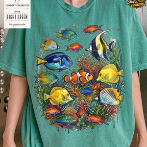 May include: Light green Comfort Colors tee featuring a vibrant watercolor illustration of various colorful fish and coral. The shirt includes the text "COMFORT COLORS TEE", "COLOR: LIGHT GREEN", and "lunagraphicscustom".
