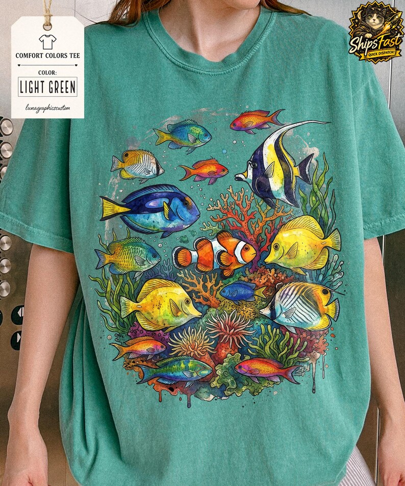 May include: Light green Comfort Colors tee featuring a vibrant watercolor illustration of various colorful fish and coral. The shirt includes the text "COMFORT COLORS TEE", "COLOR: LIGHT GREEN", and "lunagraphicscustom".