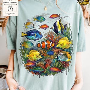 May include: A light blue Comfort Colors tee featuring a vibrant underwater scene. The design showcases colorful fish, coral, and aquatic plants. The shirt includes the text "BAY" and "lunagraphiccustom".