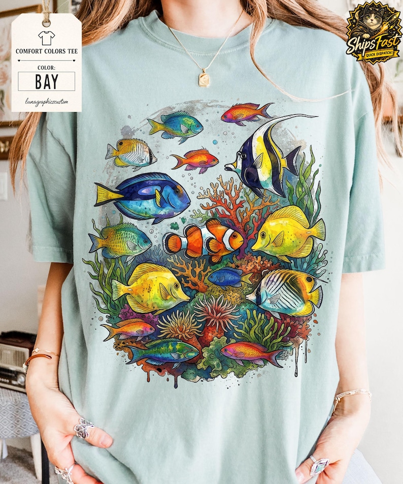May include: A light blue Comfort Colors tee featuring a vibrant underwater scene. The design showcases colorful fish, coral, and aquatic plants. The shirt includes the text "BAY" and "lunagraphiccustom".