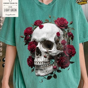 May include: Light green comfort colors tee featuring a detailed skull design encircled by vibrant red roses. The shirt includes the text "LIGHT GREEN" and "lunagraphiccustom" on a tag. The design is centered on the shirt.
