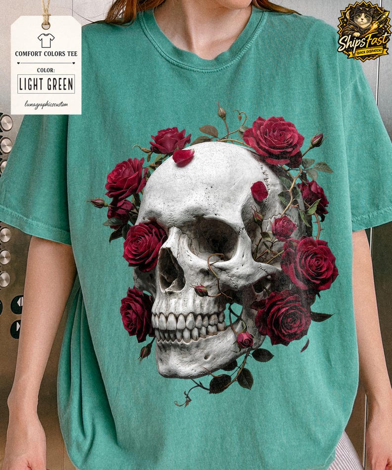 May include: Light green comfort colors tee featuring a detailed skull design encircled by vibrant red roses. The shirt includes the text "LIGHT GREEN" and "lunagraphiccustom" on a tag. The design is centered on the shirt.
