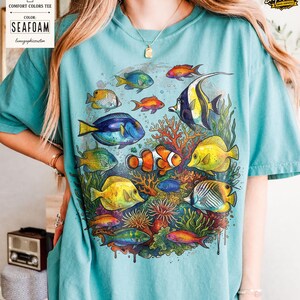May include: Seafoam-colored Comfort Colors tee featuring a vibrant illustration of various colorful fish swimming around coral. The design includes a clownfish, yellow tangs, and other tropical fish. The shirt also has the text "Seafoam" and "lunagraphiccustom".