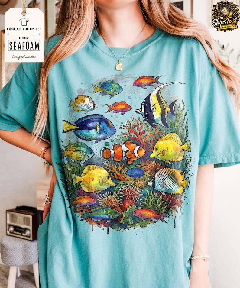 May include: Seafoam-colored Comfort Colors tee featuring a vibrant illustration of various colorful fish swimming around coral. The design includes a clownfish, yellow tangs, and other tropical fish. The shirt also has the text "Seafoam" and "lunagraphiccustom".