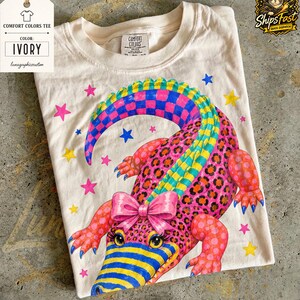 Könnte beinhalten: Elfenbeinfarbenes Comfort Colors T-Shirt mit einem farbenfrohen Alligatorendesign. Der Alligator hat einen rosa Körper mit Leopardenflecken, einen Regenbogen-Karoschwanz und eine rosa Schleife. Das Shirt hat auch rosa, blaue und gelbe Sterne.