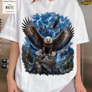 May include: White Comfort Colors tee featuring a detailed graphic of a bald eagle with outstretched wings, perched on a rocky outcrop with its young. The background includes a mountain range, waterfall, and lightning.