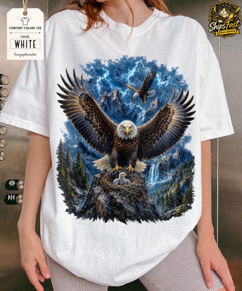 May include: White Comfort Colors tee featuring a detailed graphic of a bald eagle with outstretched wings, perched on a rocky outcrop with its young. The background includes a mountain range, waterfall, and lightning.