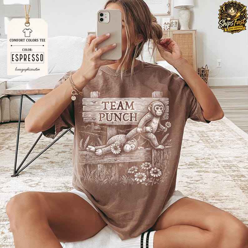 May include: Espresso-coloured comfort colours tee featuring a graphic of two monkeys on a wooden fence, with the text "TEAM PUNCH". The design includes flowers and wheat stalks. The shirt has a relaxed fit.