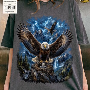 May include: Dark gray Comfort Colors tee featuring a detailed graphic of a bald eagle with outstretched wings, set against a backdrop of mountains, lightning, and a waterfall. The eagle is perched on a nest with a baby eagle. The tag reads "Pepper".