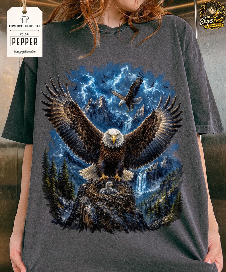 May include: Dark gray Comfort Colors tee featuring a detailed graphic of a bald eagle with outstretched wings, set against a backdrop of mountains, lightning, and a waterfall. The eagle is perched on a nest with a baby eagle. The tag reads "Pepper".