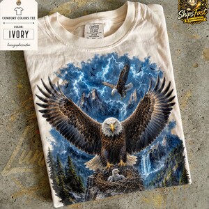 May include: Ivory Comfort Colors tee featuring a detailed graphic of a bald eagle with outstretched wings, a nest with an eaglet, and a backdrop of mountains, waterfalls, and lightning. The shirt has a tag that says "Comfort Colors Tee" and "Ivory".