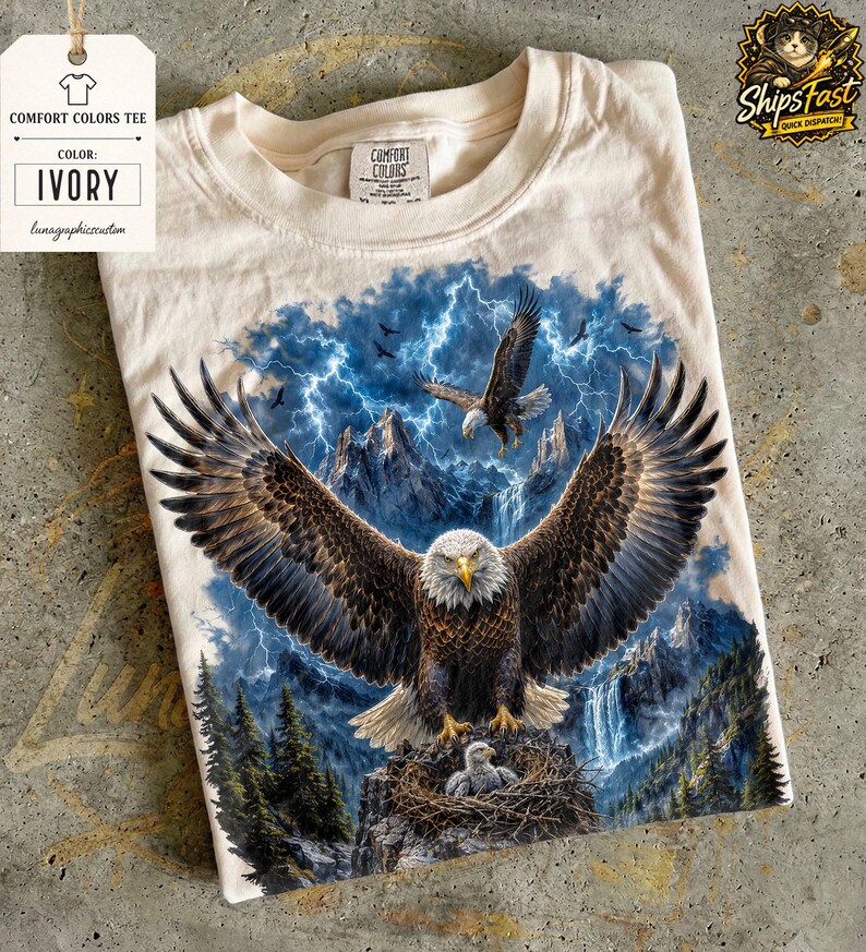 May include: Ivory Comfort Colors tee featuring a detailed graphic of a bald eagle with outstretched wings, a nest with an eaglet, and a backdrop of mountains, waterfalls, and lightning. The shirt has a tag that says "Comfort Colors Tee" and "Ivory".