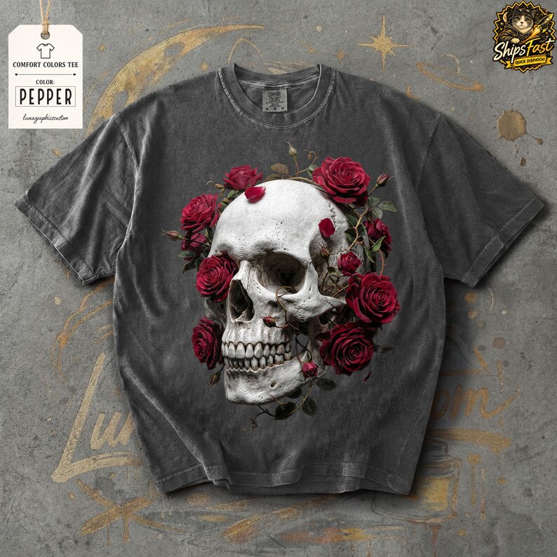 May include: A dark gray Comfort Colors tee featuring a white skull encircled by red roses. The shirt has a vintage look, with the text "Pepper" on a tag. The design is centered on the front of the shirt.