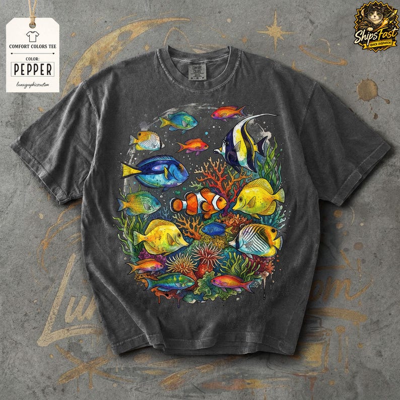 May include: Dark gray Comfort Colors tee featuring a colorful illustration of various fish species, including clownfish, angelfish, and others, swimming around coral. The shirt has the text "PEPPER" on a tag.