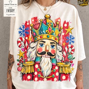 Può includere: Una t-shirt color avorio con una grafica vivace di uno schiaccianoci che indossa una corona blu e oro. Il disegno include bastoncini di zucchero rossi e bianchi, stelle gialle, fiocchi di neve blu e piccoli ornamenti su sfondo a pennellate.