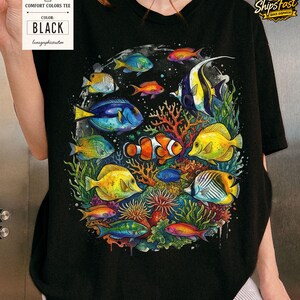 May include: Black Comfort Colors tee featuring a vibrant, colorful illustration of various tropical fish swimming around coral and seaweed. The shirt includes a tag that says "BLACK".